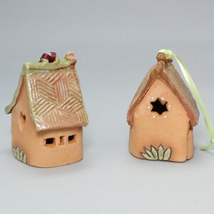 May include: Two small, hand-crafted clay houses with a rustic, cottage-style design. One house has a heart-shaped window and the other has a star-shaped window. Both houses have a green roof and are decorated with brown and white details.