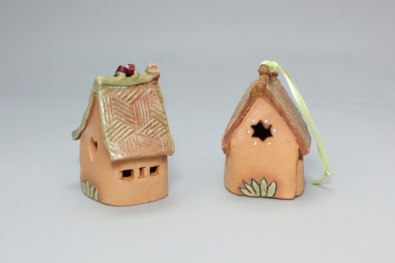 May include: Two small, hand-crafted clay houses with a rustic, cottage-style design. One house has a heart-shaped window and the other has a star-shaped window. Both houses have a green roof and are decorated with brown and white details.