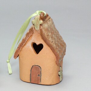 May include: A small, terracotta-colored ceramic house ornament with a heart-shaped window, a cross on the roof, and a green ribbon for hanging. The house has a brown door and a cross on the side.