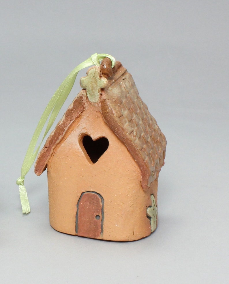 May include: A small, terracotta-colored ceramic house ornament with a heart-shaped window, a cross on the roof, and a green ribbon for hanging. The house has a brown door and a cross on the side.