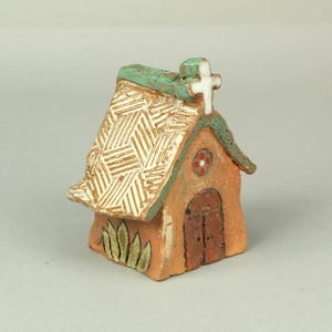 May include: A small, hand-crafted ceramic church with a white cross on top, a green roof, and a brown door. The church has a textured roof and a flower design on the front.