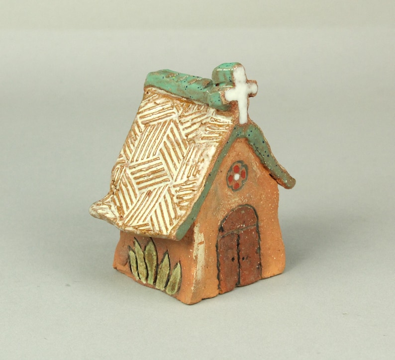 May include: A small, hand-crafted ceramic church with a white cross on top, a green roof, and a brown door. The church has a textured roof and a flower design on the front.