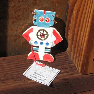 May include: A handmade ceramic robot ornament with red, white, and blue paint. The robot has a round head with blue eyes and a gear on its chest. The ornament is hanging from a red string and has a tag that reads "Handmade by Arizona Artist Karlene Voepel".