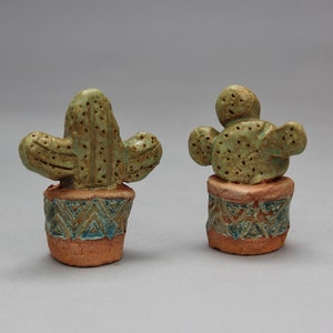 May include: Two small ceramic cactus plants in pots with a geometric pattern. The cactus on the left is tall and slender with two arms, while the cactus on the right is rounder with multiple arms.