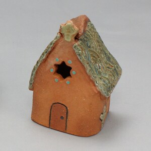 May include: A small, hand-crafted ceramic house with a brown clay body and a green and brown glazed roof. The house has a small door and a window with a star-shaped opening and blue accents.
