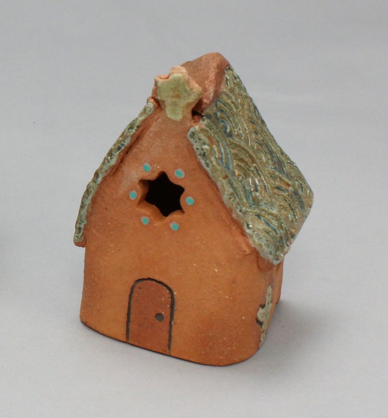 May include: A small, hand-crafted ceramic house with a brown clay body and a green and brown glazed roof. The house has a small door and a window with a star-shaped opening and blue accents.
