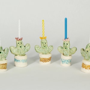 May include: Five ceramic cactus candle holders with different colored candles. Each cactus has a happy face and is wearing a small hat. The cactus are in small pots with different colored designs.
