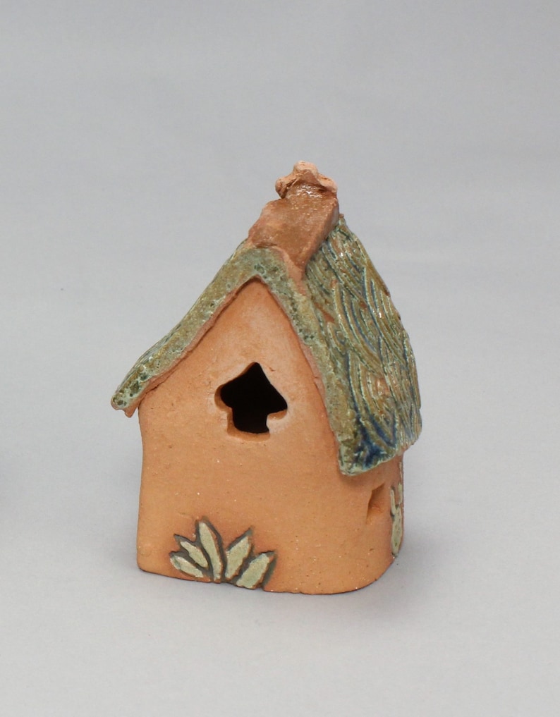 May include: A small, hand-crafted ceramic house with a green and brown roof, a small window, and a decorative design on the front.