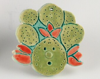 May include: A ceramic wall hanging with a green and blue floral design. The design features a central flower with a face and a ring of smaller flowers around it.