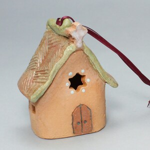 May include: A small, hand-crafted ceramic house ornament with a thatched roof, a star on top, and a door. The ornament is painted in shades of brown, green, and white, and has a burgundy ribbon attached for hanging.