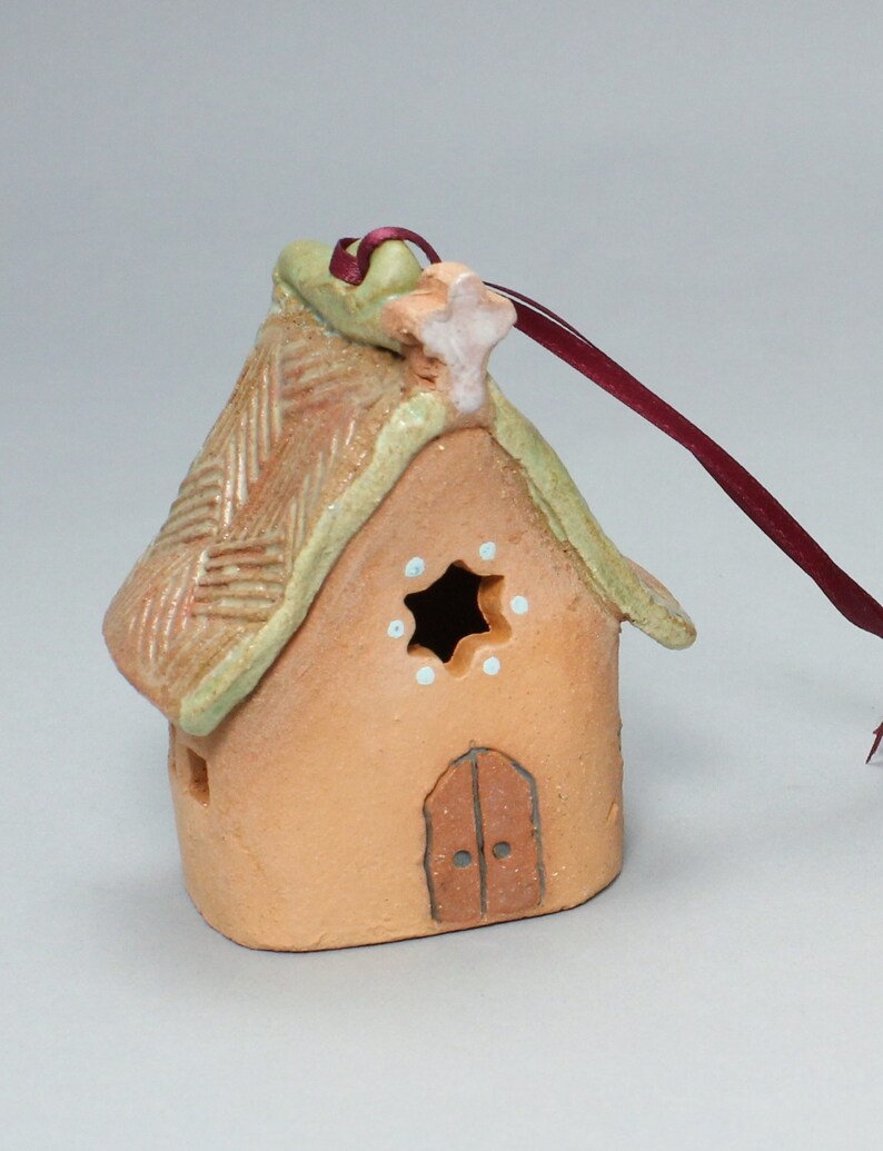 May include: A small, hand-crafted ceramic house ornament with a thatched roof, a star on top, and a door. The ornament is painted in shades of brown, green, and white, and has a burgundy ribbon attached for hanging.
