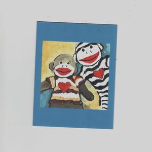 May include: A colorful painting of two sock monkey characters. One is brown with a striped shirt and a red heart, the other is black and white striped with a red heart. Both have red mouths and are set against a yellow and blue background.