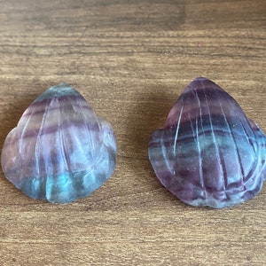 May include: Two carved fluorite gemstone shells. The shells are a mix of purple, blue, and clear colours. The shells are on a wooden surface. The shells have a detailed carved design.