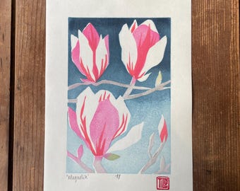 May include: A rectangular art print on off-white paper featuring three pink and white magnolia blossoms on a branch against a dark blue gradient background. The word Magnolia is written in the bottom left, with a red square stamp in the bottom right.