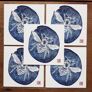 May include: Five square art prints featuring a detailed white bee illustration set against a dark blue tree ring background. Each print has a red square stamp in the corner. The prints are arranged on a wooden surface.