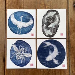 May include: Four square art prints featuring white ink illustrations on blue and black backgrounds. The prints depict a crane in flight, three koi fish, a bee, and a single koi fish, all set within circular wood grain textures with a small red stamp.