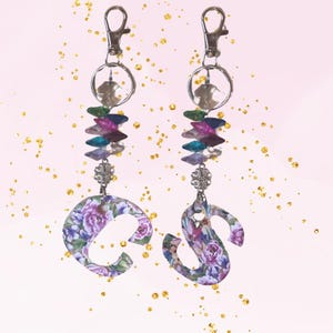 May include: Two keychains with floral patterned letter charms, silver key rings, and crystal beads in shades of purple, pink, blue, and green.