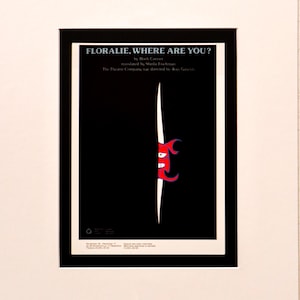 May include: A framed poster with the title "FLORALIE, WHERE ARE YOU?" in white text against a black background. A red cartoon face peeks out from a white vertical line. The poster is in a white mount and black frame.