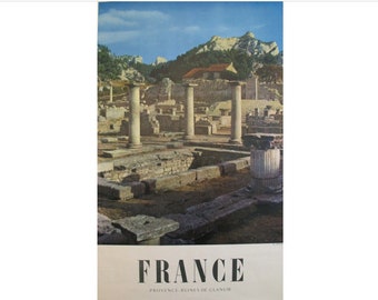 May include: A vintage travel poster featuring a photograph of the ruins of Glanum in Provence, France. The poster features a black and white image of the ruins with a mountain range in the background. The text "FRANCE" is printed in bold black letters at the bottom of the poster, with the words "PROVENCE, RUINES DE GLANUM" printed in smaller black letters below.