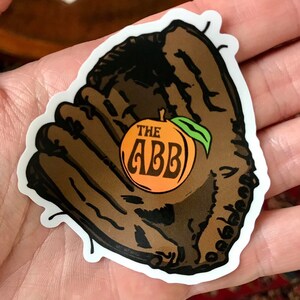 May include: A brown leather baseball glove with a peach inside. The peach has the text "THE ABB" on it.