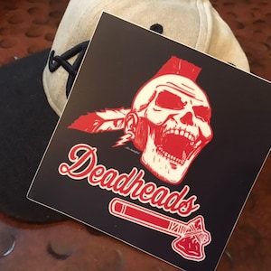 May include: A red and white sticker with a skull wearing a mohawk and a feather headdress. The skull is yelling and has a red and white tomahawk below it. The text "Deadheads" is written in a red cursive font.