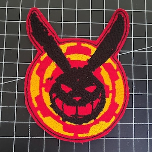Könnte beinhalten: Gestickter Aufnäher mit einem stilisierten, grinsenden Kaninchengesicht mit großen Ohren. Das Design verwendet die Farben Schwarz, Rot und Gelb. Das Kaninchengesicht ist vor einem roten und gelben kreisförmigen Hintergrund platziert.