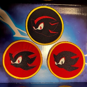 Könnte beinhalten: Drei runde, gestickte Aufnäher mit der Figur Shadow aus der Sonic the Hedgehog-Serie. Jeder Aufnäher zeigt eine schwarze Silhouette von Shadows Gesicht, umrandet in Rot und Gelb. Der Hintergrund ist ein blauer Farbverlauf.