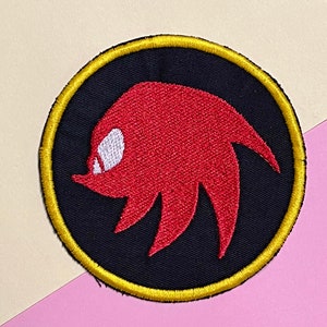 Könnte beinhalten: Gestickter Aufnäher mit einem roten Charakterdesign und einem weißen Auge auf schwarzem Hintergrund. Der Aufnäher hat einen gelben Rand und ist kreisförmig. Das Design ist von einem bekannten Videospielcharakter inspiriert.
