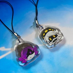 Peut inclure: Deux breloques en acrylique transparent avec des cordons noirs. L'une représente un personnage aux cheveux violets avec un bonnet gris. L'autre présente un personnage aux cheveux blancs avec un motif jaune et noir. Les deux ont un fermoir argenté.