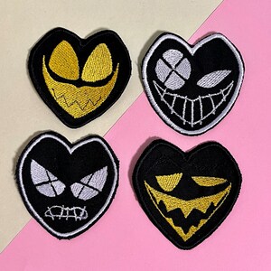 May include: Four black heart-shaped embroidered patches with different facial expressions. Two have gold features, and two have white features. The patches are on a pink and beige background.