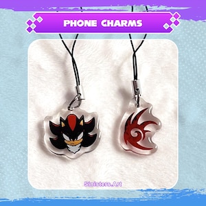 Può includere: Due ciondoli per telefono in acrilico trasparente con cordini neri. Un ciondolo presenta un design di personaggio nero e rosso, l'altro un design tribale rosso. Il testo "PHONE CHARMS" è in uno striscione viola in alto.