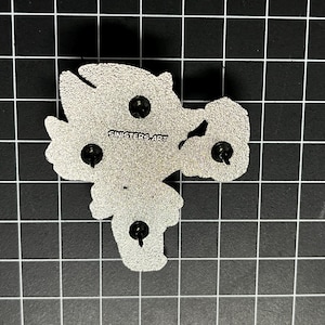 May include: The back of a silver-colored enamel pin with three pin backs. The pin is in the shape of a cartoon character. The text "SINISTERS ART" is printed on the pin.