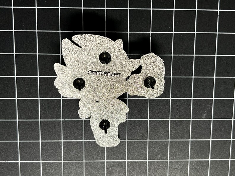 May include: The back of a silver-colored enamel pin with three pin backs. The pin is in the shape of a cartoon character. The text "SINISTERS ART" is printed on the pin.