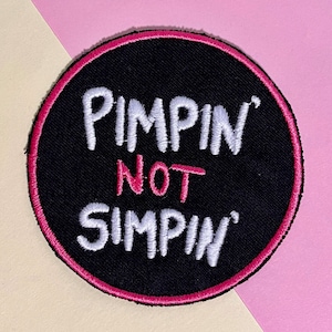 Könnte beinhalten: Gestickter schwarzer Aufnäher mit den Worten "PIMPIN NOT SIMPIN" in weißer und rosa Schrift. Der Aufnäher hat einen rosa Rand und ist vor einem rosa-cremefarbenen Hintergrund platziert.