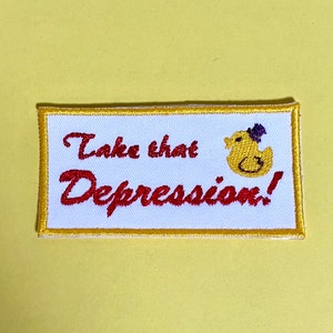 May include: Embroidered patch with the phrase "Take that Depression!" in red script. A yellow duck wearing a purple top hat is on the right. The patch has a white background with a gold border, set against a yellow backdrop.
