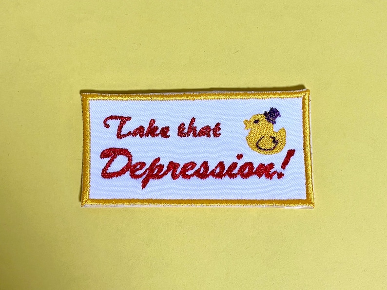 May include: Embroidered patch with the phrase "Take that Depression!" in red script. A yellow duck wearing a purple top hat is on the right. The patch has a white background with a gold border, set against a yellow backdrop.