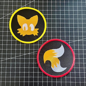 以下が含まれることがあります： ソニック・ザ・ヘッジホッグシリーズのキャラクター、テイルスの刺繍パッチ2枚。1枚は、黄と白のテイルスの顔が黒い背景に黄色の縁取りで描かれています。もう1枚は、黄と白の2本の尻尾が黒い背景に赤い縁取りで描かれています。