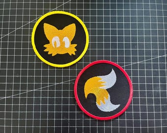 Könnte beinhalten: Zwei gestickte Aufnäher mit der Figur Tails aus der Sonic the Hedgehog-Serie. Ein Aufnäher zeigt Tails' Gesicht in Gelb und Weiß auf schwarzem Hintergrund mit gelbem Rand. Der andere zeigt seine beiden Schwänze in Gelb und Weiß auf schwarzem Hintergrund mit rotem Rand.