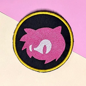 Könnte beinhalten: Runder, gestickter Aufnäher mit einem rosa Charakterdesign auf schwarzem Hintergrund, goldfarben eingefasst. Der Charakter hat ein weißes Gesicht und stachelige Haare. Geeignet für Kleidung oder Accessoires.