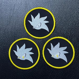 Könnte beinhalten: Drei kreisförmige, gestickte Aufnäher mit einem stilisierten silbernen Igelkopf mit gelben Augen und einem stacheligen Design. Die Aufnäher befinden sich auf einem schwarzen Hintergrund mit gelbem Rand.