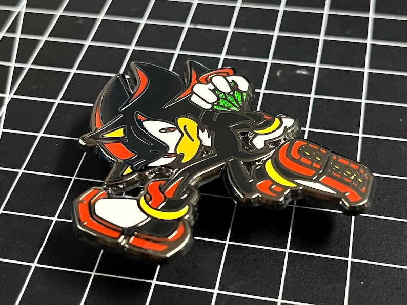 May include: A metal pin featuring the character Shadow the Hedgehog from the Sonic the Hedgehog video game series. The pin is black, red, yellow, and white, and depicts Shadow holding a green Chaos Emerald.
