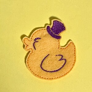 May include: A felt duck applique in light yellow, wearing a purple top hat. The duck has a stitched outline and details in purple and gold. The applique is on a light yellow background.