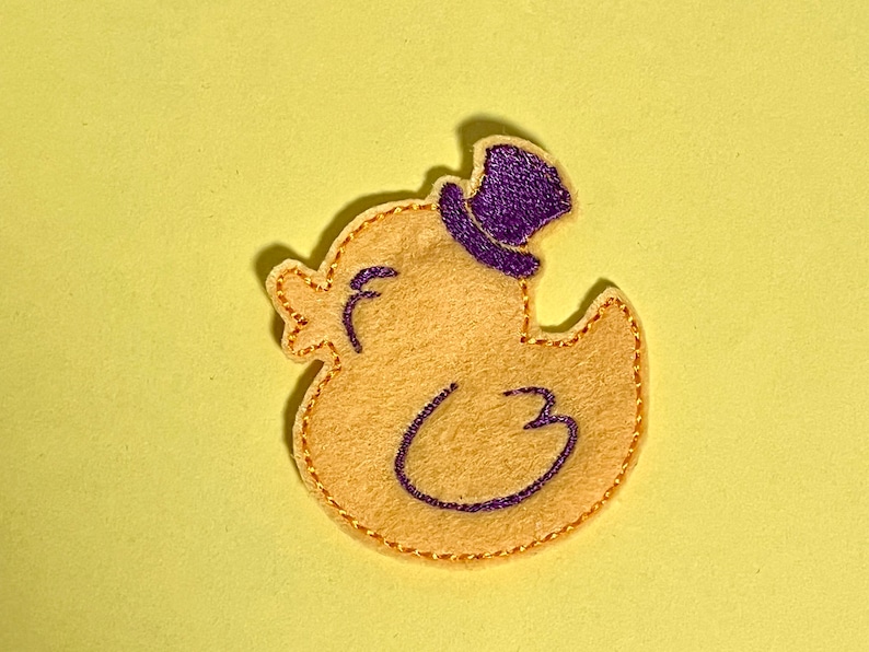 May include: A felt duck applique in light yellow, wearing a purple top hat. The duck has a stitched outline and details in purple and gold. The applique is on a light yellow background.