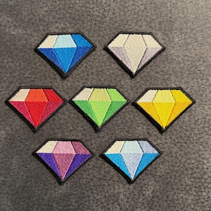 Puede incluir: Siete parches bordados en forma de diamante con bordes negros. Los parches presentan varios colores, incluyendo azul, blanco, rojo, verde, amarillo, morado y azul claro, diseñados con facetas geométricas que recuerdan a una piedra preciosa.