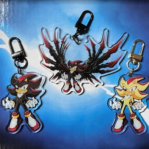 May include: Three acrylic keychains featuring characters from a video game. The keychains depict a black and red character, a character with black wings and a red and black design, and a yellow and red character. Each keychain has a black or gold-coloured clasp.