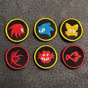 May include: Six circular embroidered patches featuring character icons from the Sonic the Hedgehog franchise. The patches include red, blue, and yellow thread designs on black backgrounds with yellow or red stitched borders.