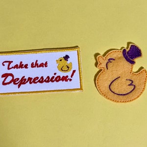 May include: Two embroidered patches on a yellow background. One rectangular patch reads "Take that Depression!" with a duck illustration. The other patch is a yellow duck wearing a purple top hat.