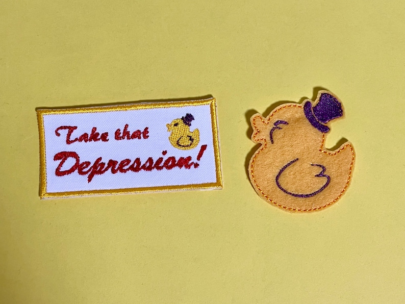 May include: Two embroidered patches on a yellow background. One rectangular patch reads "Take that Depression!" with a duck illustration. The other patch is a yellow duck wearing a purple top hat.
