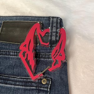 K&ouml;nnte beinhalten: Ein pinkfarbenes Acryl-Accessoire, m&ouml;glicherweise ein Schl&uuml;sselanh&auml;nger oder ein Anh&auml;nger, ist an der Ges&auml;&szlig;tasche einer blauen Jeans befestigt. Das Accessoire hat ein stilisiertes Design mit scharfen Winkeln und einer Herzform.