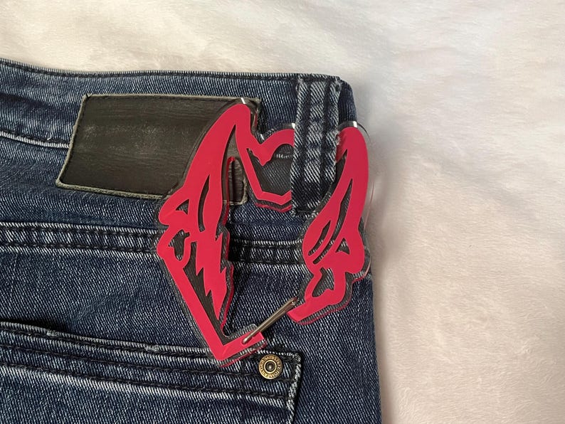 K&ouml;nnte beinhalten: Ein pinkfarbenes Acryl-Accessoire, m&ouml;glicherweise ein Schl&uuml;sselanh&auml;nger oder ein Anh&auml;nger, ist an der Ges&auml;&szlig;tasche einer blauen Jeans befestigt. Das Accessoire hat ein stilisiertes Design mit scharfen Winkeln und einer Herzform.